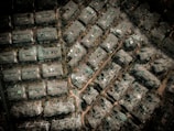 An aerial view of a densely packed urban area with numerous rectangular buildings. The structures are arranged in a grid-like pattern, surrounded by small patches of greenery and narrow pathways. The buildings appear to be residential with flat roofs and varied structural details.