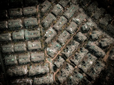 An aerial view of a densely packed urban area with numerous rectangular buildings. The structures are arranged in a grid-like pattern, surrounded by small patches of greenery and narrow pathways. The buildings appear to be residential with flat roofs and varied structural details.