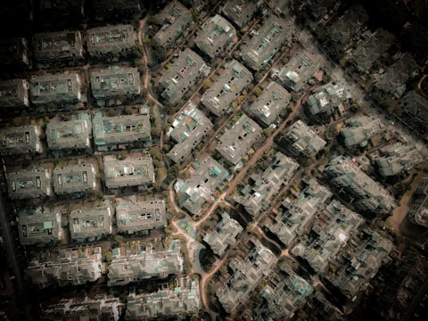 An aerial view of a densely packed urban area with numerous rectangular buildings. The structures are arranged in a grid-like pattern, surrounded by small patches of greenery and narrow pathways. The buildings appear to be residential with flat roofs and varied structural details.