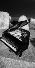 A small grand piano is placed on a rough stone surface, surrounded by large, irregularly shaped rocks. The piano's lid is open, revealing its strings and hammers. The entire scene is in black and white, giving it a timeless and artistic feel.