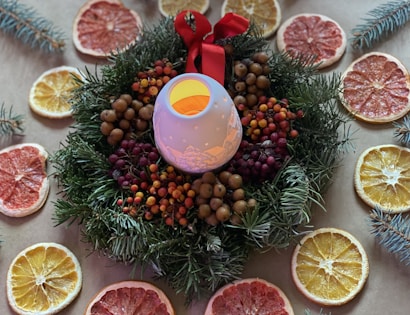 A decorative arrangement featuring a candle holder in the center with a red bow, surrounded by a wreath made of evergreen branches and various berries. Slice of dried citrus fruits, including oranges and grapefruits, are placed in a circle around the wreath on a brown surface.