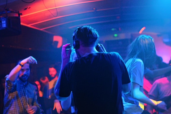 A dimly lit nightclub with colorful lighting where several people are dancing and enjoying the music. A person with headphones stands in the center, possibly a DJ, while others move around energetically.
