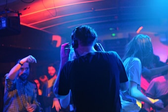 A dimly lit nightclub with colorful lighting where several people are dancing and enjoying the music. A person with headphones stands in the center, possibly a DJ, while others move around energetically.
