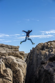man in black jacket and blue denim jeans jumping on brown rocky mountain during daytime