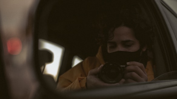 A person is taking a photo of themselves using a camera while looking into a car's side mirror. The person has curly hair and is wearing a mask and a jacket. The viewpoint is intimate, suggesting introspection or contemplation.