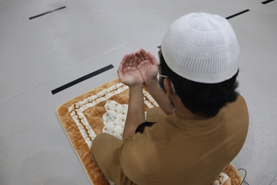 A person wearing a white cap and brown clothing is seated on a plush prayer mat with hands raised in a gesture of prayer or supplication. The setting appears to be indoors with a light-colored floor and black lines visible on the surface.