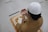 A person wearing a white cap and brown clothing is seated on a plush prayer mat with hands raised in a gesture of prayer or supplication. The setting appears to be indoors with a light-colored floor and black lines visible on the surface.