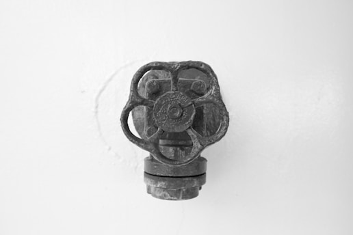 A metallic valve with a four-pronged, circular handle protrudes from a flat, plain wall. The surface of the valve is textured, indicating age or wear, and it contrasts against the smooth background.