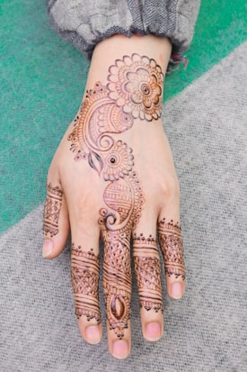 A detailed henna design is drawn on the back of a hand, featuring intricate floral and paisley patterns. The artwork extends from the wrist to the fingers, showcasing elaborate motifs and delicate line work.