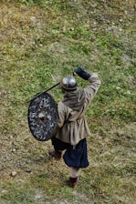 A person dressed in medieval armor is holding a round shield and appears to be in mid-stride on a grassy terrain. The helmet is metallic, and the shield shows signs of wear. The person is outfitted in a tunic, dark pants, and sturdy shoes.