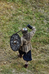 A person dressed in medieval armor is holding a round shield and appears to be in mid-stride on a grassy terrain. The helmet is metallic, and the shield shows signs of wear. The person is outfitted in a tunic, dark pants, and sturdy shoes.