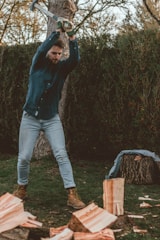 A person is chopping wood with an axe in a wooded area, wearing a blue checkered shirt, light jeans, and brown work boots. Logs and wood chips are scattered on the ground, and there is a tree in the background.