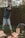 A person is chopping wood with an axe in a wooded area, wearing a blue checkered shirt, light jeans, and brown work boots. Logs and wood chips are scattered on the ground, and there is a tree in the background.