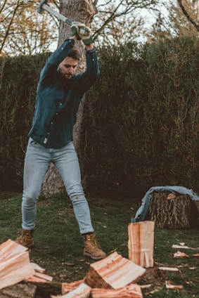 A person is chopping wood with an axe in a wooded area, wearing a blue checkered shirt, light jeans, and brown work boots. Logs and wood chips are scattered on the ground, and there is a tree in the background.