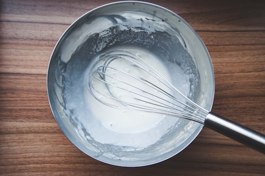 A metal mixing bowl containing a creamy white mixture sits on a wooden surface. A stainless steel whisk is partially submerged in the mixture, suggesting preparation of a recipe. The interior of the bowl shows traces of the mixture spread around the edges.