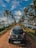 A black Hyundai Eon car is parked on a narrow dirt road flanked by lush green palm trees. A serene water body is on the right side, reflecting the palm trees and clear blue sky, which is dotted with white clouds.