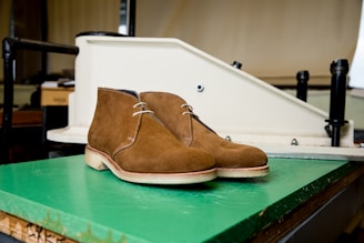 A pair of brown suede boots with white soles is placed on a green floor in front of industrial equipment. The background includes a few dark mechanical parts and a beige wall.