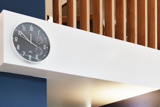 A modern interior features a large, white horizontal beam with a black analog clock mounted on it. The clock displays numbers in a clear, traditional style. Behind the beam, vertical wooden slats add a textured architectural element. The overall setting conveys a minimalist and contemporary design aesthetic.