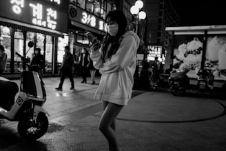 A nighttime city street scene features a person wearing a mask and a hooded sweatshirt, looking at a smartphone. There are several people in the background, walking along the sidewalk. Bright signs and storefronts illuminate the area, casting light and shadows across the scene. A stationary scooter is visible, parked nearby.