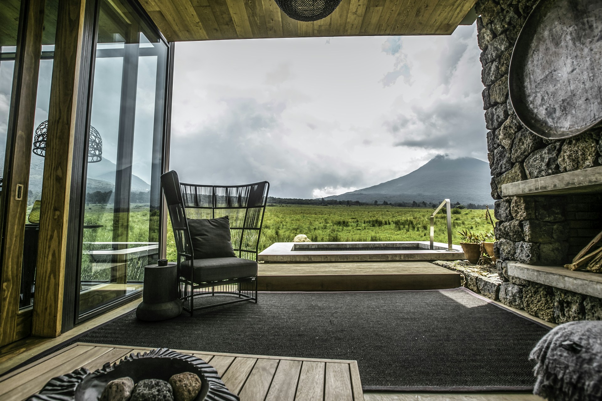 A cozy living space with modern furniture, large glass windows overlooking a scenic view of lush, verdant fields and a towering mountain under a cloudy sky. The room features a stone wall, wooden elements, and a stylish black chair with a cushion. A few planters and an indoor rug add to the aesthetic.