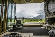 A cozy living space with modern furniture, large glass windows overlooking a scenic view of lush, verdant fields and a towering mountain under a cloudy sky. The room features a stone wall, wooden elements, and a stylish black chair with a cushion. A few planters and an indoor rug add to the aesthetic.