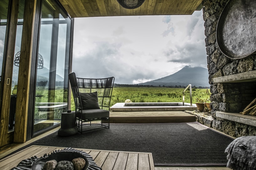 A cozy living space with modern furniture, large glass windows overlooking a scenic view of lush, verdant fields and a towering mountain under a cloudy sky. The room features a stone wall, wooden elements, and a stylish black chair with a cushion. A few planters and an indoor rug add to the aesthetic.