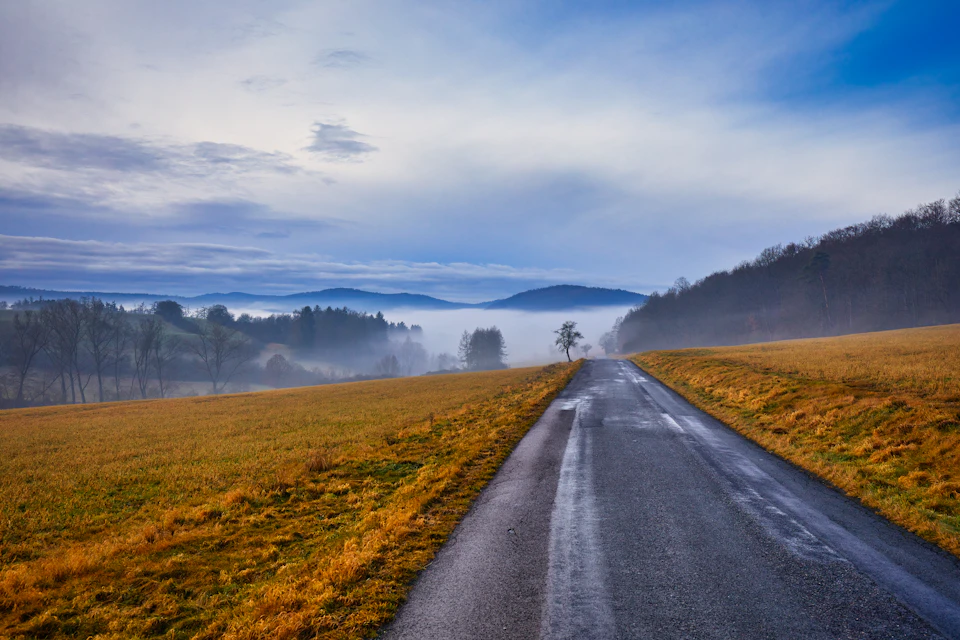 The image depicts a serene rural landscape with an asphalt road cutting through it, flanked by lush green fields on both sides. The sky above is filled with clouds, casting a soft light over the scene and creating a sense of tranquility. In the distance, rolling hills can be seen under the cloudy sky, adding depth to the image. A lone tree stands in the foreground, its presence enhancing the natural beauty of the landscape.