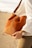 A person holding a stylish brown leather handbag, complemented by a white blouse and beige trousers. The person is wearing a gold wristwatch and a ring, with neatly groomed nails painted burgundy. The background is soft and blurred, suggesting an outdoor setting with sand.