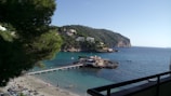 A scenic coastal area with clear blue water and a small beach. A wooden pier leads to a rocky outcrop with a building, surrounded by lush green hills. People are relaxing on the sandy beach with umbrellas scattered around. Residential houses can be seen on the hill in the background.