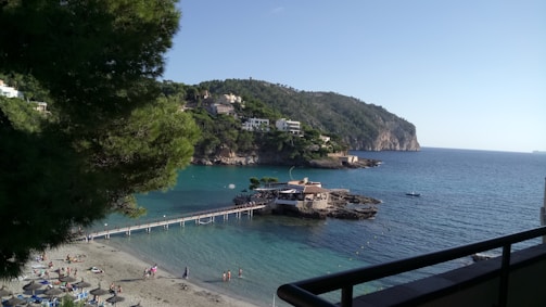 A scenic coastal area with clear blue water and a small beach. A wooden pier leads to a rocky outcrop with a building, surrounded by lush green hills. People are relaxing on the sandy beach with umbrellas scattered around. Residential houses can be seen on the hill in the background.