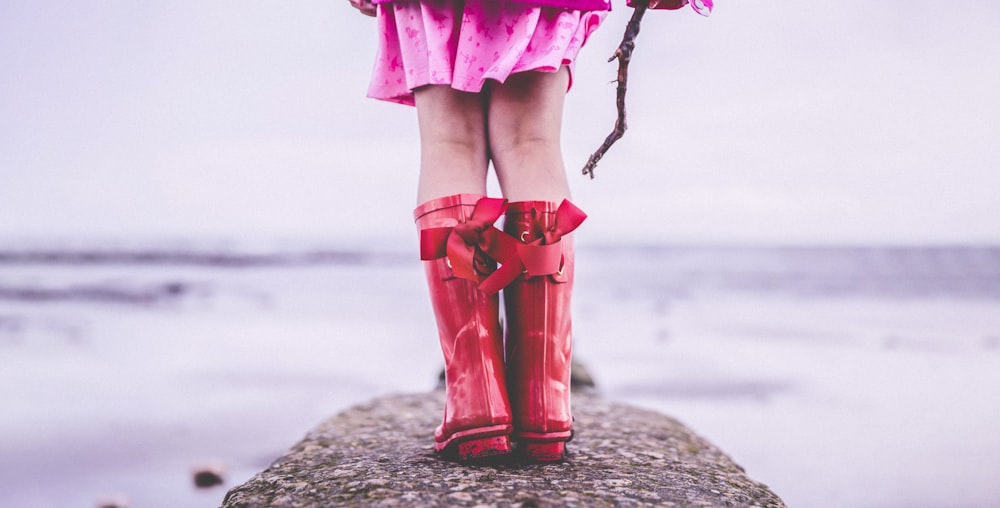 Mujer en vestido rosa y botas rojas de pie en la orilla de la playa durante el día