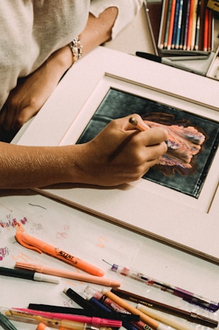 A person is engaged in drawing or coloring on a framed piece of art, surrounded by a variety of colored pencils, markers, and writing utensils. The hand holding a colored pencil focuses on adding details to the artwork. There is a sketch or painting in progress, with vibrant hues. The workspace suggests creativity and artistic activity.