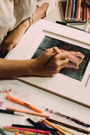 A person is engaged in drawing or coloring on a framed piece of art, surrounded by a variety of colored pencils, markers, and writing utensils. The hand holding a colored pencil focuses on adding details to the artwork. There is a sketch or painting in progress, with vibrant hues. The workspace suggests creativity and artistic activity.