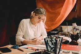 A thoughtful woman writing at a desk surrounded by forensic science materials.