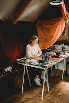 A person is seated at a cluttered desk filled with various art supplies, notebooks, and electronic devices. The room has a rustic feel with wooden floors and exposed beams. Dramatic lighting casts warm shadows, with vibrant red and orange fabrics draped in the background.