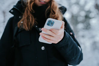 A person with long hair dressed in a dark winter jacket is holding a smartphone. The background is snowy, suggesting a cold, outdoor environment.