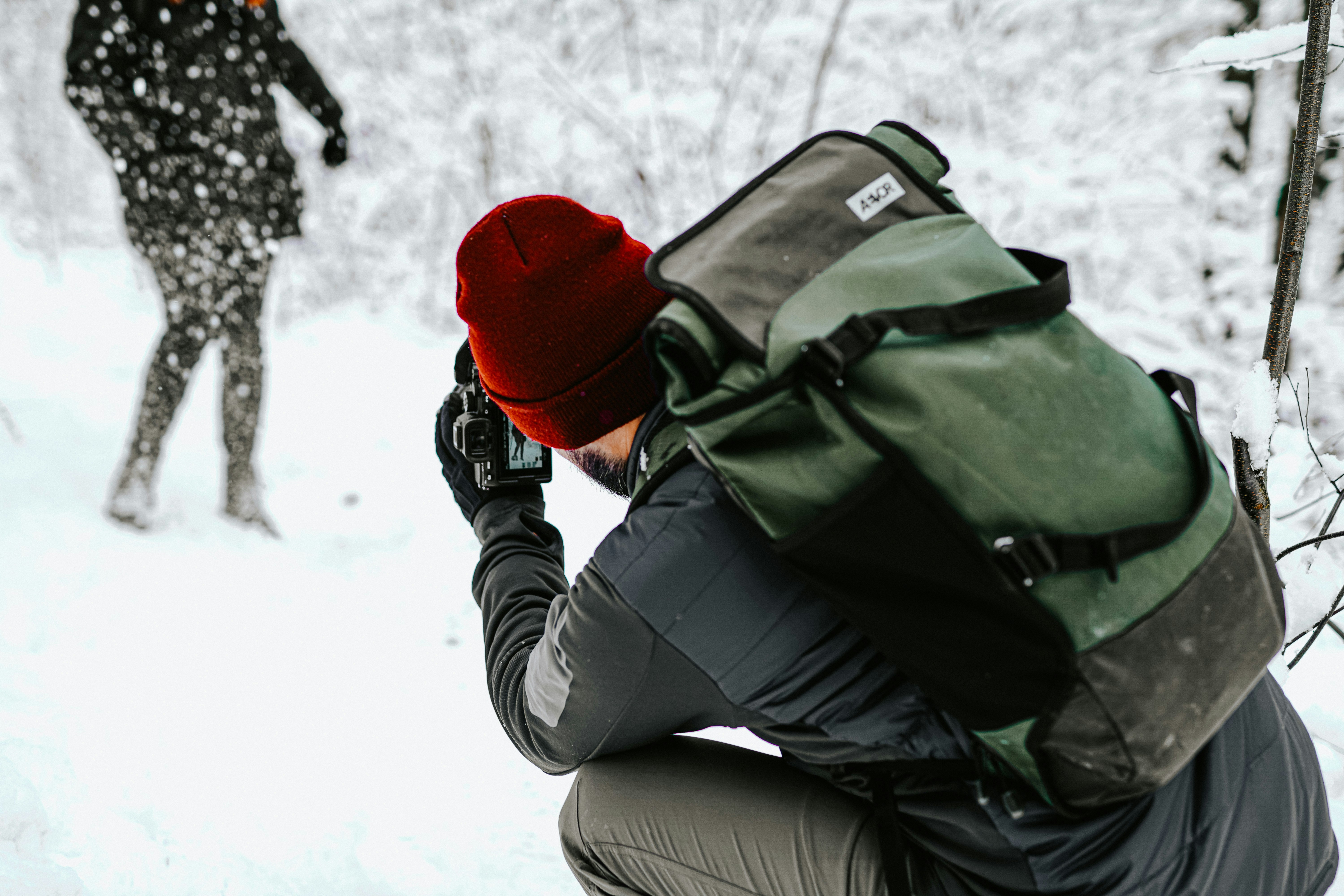 woman in black jacket and red knit cap holding black dslr camera, A photographer in the snowy landscape with a beenie and a rucksack taking pictures from a model dancing in the snow. A happy work and travel working place. Having an office outside is the best thing in the world. 