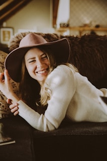 A person is smiling brightly while lounging on a couch. They are wearing a wide-brimmed hat and a cozy sweater. The background features soft, earthy tones with blurred details of a cozy room setting.