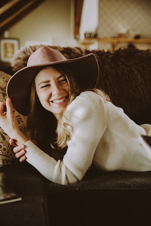A person is smiling brightly while lounging on a couch. They are wearing a wide-brimmed hat and a cozy sweater. The background features soft, earthy tones with blurred details of a cozy room setting.