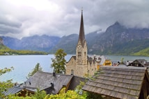A picturesque lakeside village features a prominent stone church with a tall spire in the foreground. Surrounding the church are traditional buildings with sloped roofs, set against a backdrop of a large lake and mountainous landscape. The sky is overcast, with grey clouds creating a serene and somewhat dramatic atmosphere.