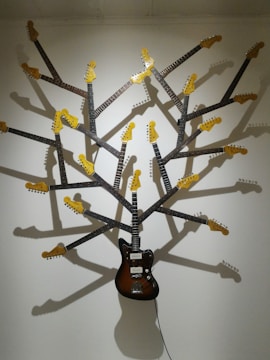 A wall-mounted art installation made from multiple guitar necks and headstocks arranged in a radial pattern, resembling a tree with branches. The central piece is a full-bodied electric guitar with a dark brown finish. Shadows cast by the guitar necks enhance the tree-like appearance on the white wall.