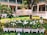 A sign labeled 'Binarn Pathom Residence' is surrounded by white orchids planted in decorative white picket-fenced planters. The area is lush with green grass and a few orange flowers scattered about. Behind the sign, there is a building with a colonial-style architecture featuring white walls and green shutters. A tree with vibrant green leaves and orange flowers is present on the left side.