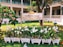 A sign labeled 'Binarn Pathom Residence' is surrounded by white orchids planted in decorative white picket-fenced planters. The area is lush with green grass and a few orange flowers scattered about. Behind the sign, there is a building with a colonial-style architecture featuring white walls and green shutters. A tree with vibrant green leaves and orange flowers is present on the left side.