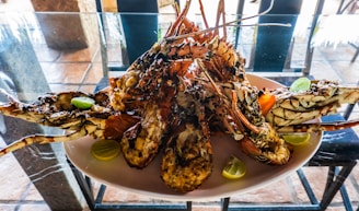A large serving of grilled lobsters on a white plate, garnished with lime wedges. The lobsters are arranged aesthetically with some cut open to reveal cooked meat. The setting appears to be a dining table with a glass top, and there are blurred elements of an outdoor backdrop.