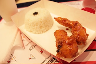 A serving of white rice and two pieces of glazed fried chicken placed in a rectangular cardboard tray. The rice has a small garnish on top, and the tray is set on a paper placemat, with parts of a napkin and cup visible in the background.