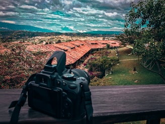 A DSLR camera is positioned on a wooden railing overlooking a scenic landscape. The view includes red-roofed buildings surrounded by lush greenery and trees. A vast expanse of hills and a cloudy sky can be seen in the background.
