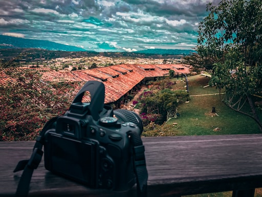 A DSLR camera is positioned on a wooden railing overlooking a scenic landscape. The view includes red-roofed buildings surrounded by lush greenery and trees. A vast expanse of hills and a cloudy sky can be seen in the background.