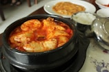 A steaming bowl of Korean stew with large chunks of tofu immersed in a spicy red broth is presented in a black stone pot. Accompanying the stew, there is a small bowl of white rice and what appears to be a Korean pancake on a separate white plate in the background.