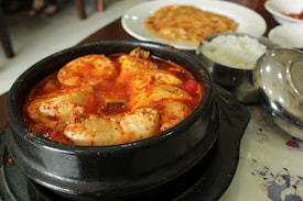 A steaming bowl of Korean stew with large chunks of tofu immersed in a spicy red broth is presented in a black stone pot. Accompanying the stew, there is a small bowl of white rice and what appears to be a Korean pancake on a separate white plate in the background.