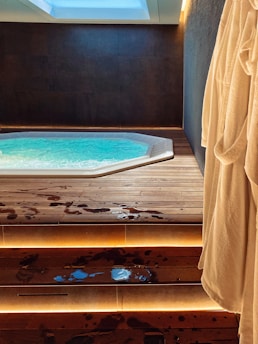 A modern indoor hot tub with bubbling water is set in a dark, minimalist room. Wooden steps lead up to the tub, with water droplets scattered across the surface. Soft, indirect lighting highlights the steps, and a plush bathrobe hangs on the right side of the image.